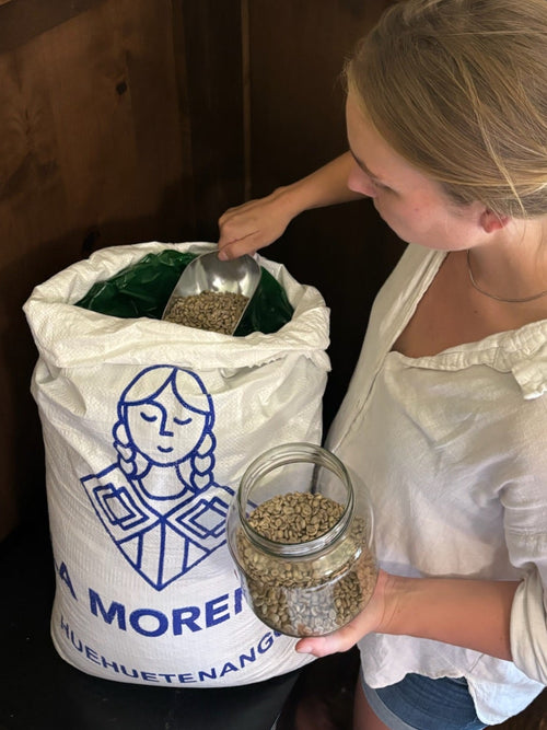 A woman in a white blouse and denim shorts stands in a corner of a room with dark wood paneling, holding a glass jar filled with dried green coffee beans while reaching into a large white sack labeled 'LA MORENA' with a metal scoop.