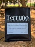 A black bag of Terruño Coffee Roasters Huchuetenango Guatemala coffee stands on a stone outdoors with a blurred forest background.