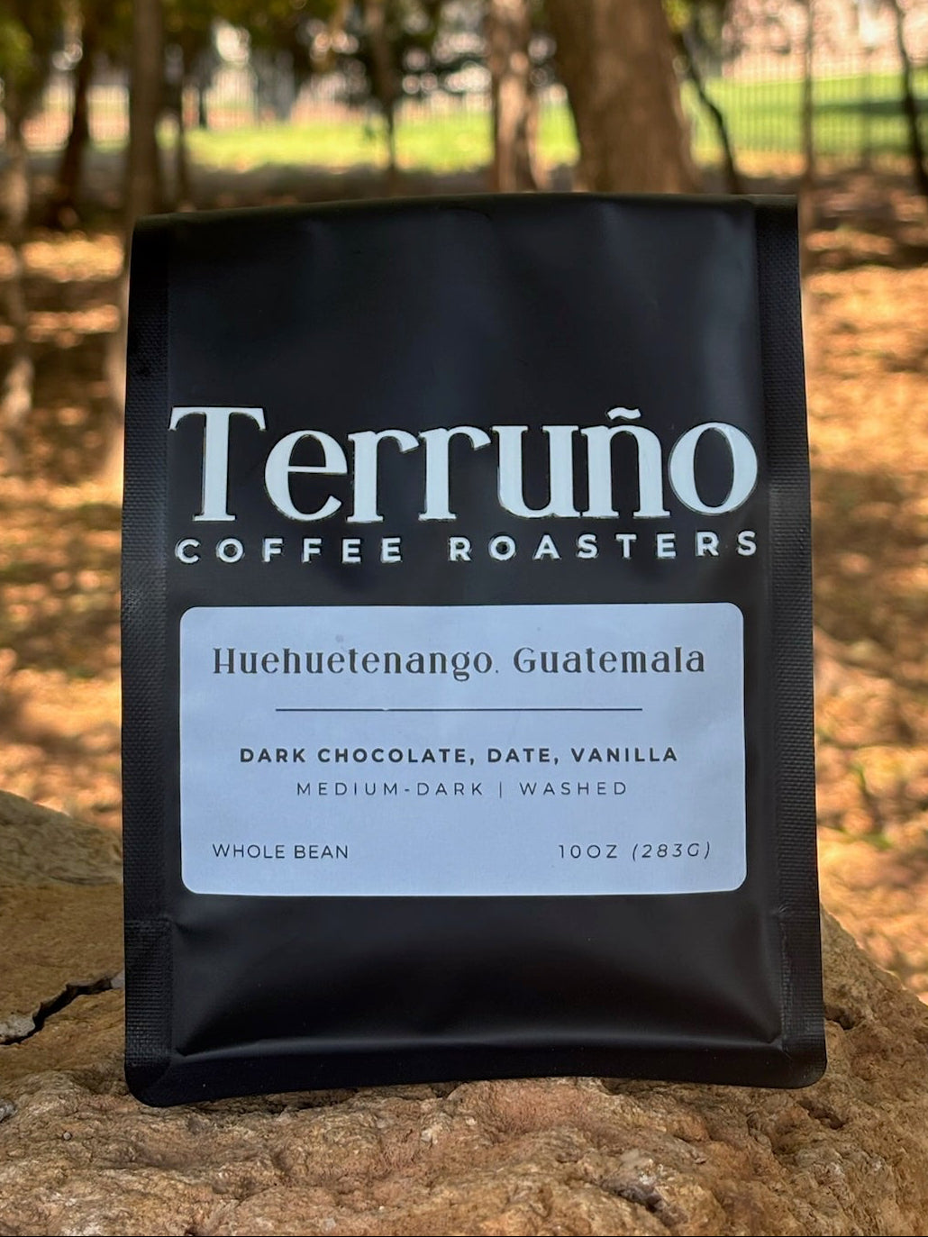 A black bag of Terruño Coffee Roasters Huchuetenango Guatemala coffee stands on a stone outdoors with a blurred forest background.